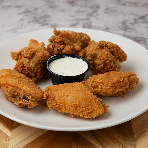 Six crispy fried chicken wings and a small black cup of ranch dressing on a white plate.